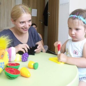 Dziewczynka z implantem ślimakowym siedzi przy stole i przygląda się kolorowym zabawkom.